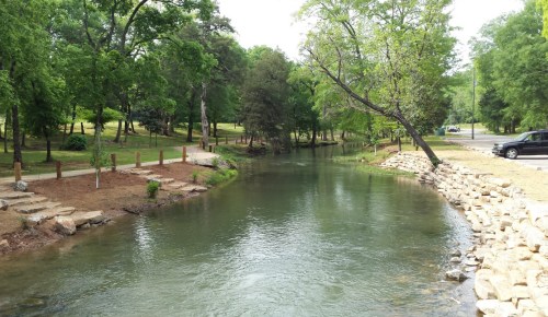 A serene river flows through a lush green park, lined with trees and rocky banks, with a car parked nearby.