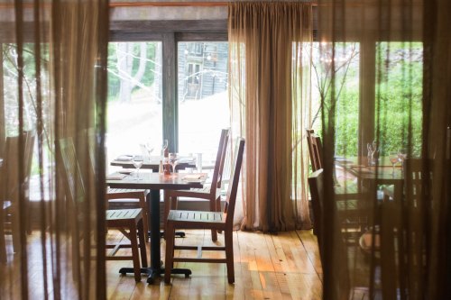 A cozy restaurant interior with wooden tables and chairs, partially obscured by sheer curtains, overlooking greenery.