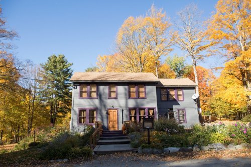 A two-story blue house with purple windows surrounded by colorful autumn trees and a garden.