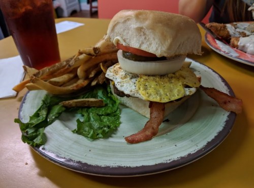 A burger with bacon, fried egg, lettuce, tomato, and onion, served with fries on a plate.
