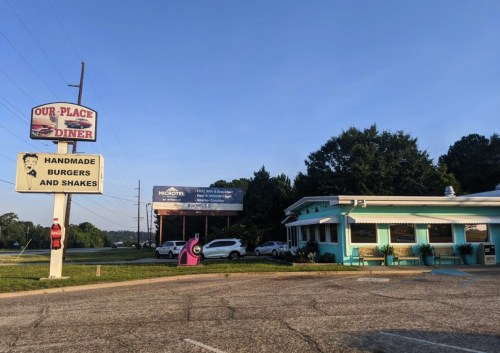A retro diner with a sign for handmade burgers and shakes, surrounded by trees and a clear blue sky.