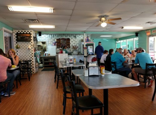 A casual diner interior with tables, patrons eating, and a visible kitchen area in the background.