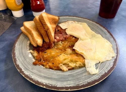 A plate of breakfast featuring hash browns, two fried eggs, bacon, and toast, with condiments in the background.