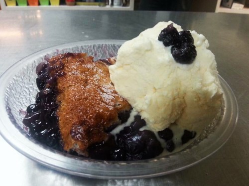 A bowl of blueberry cobbler topped with a scoop of vanilla ice cream, served on a silver countertop.