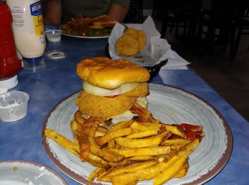 A plate with a stacked fried chicken sandwich, pickles, and a side of crispy fries, with condiments in the background.