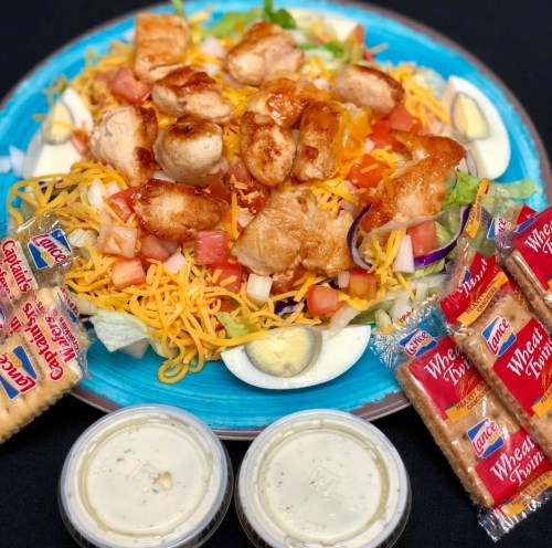 A colorful salad topped with grilled chicken, cheese, and vegetables, accompanied by crackers and two containers of dressing.