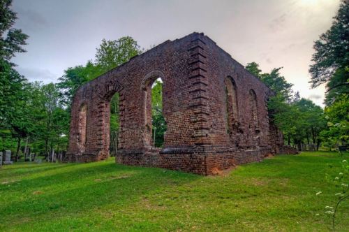 Ruins of a brick structure surrounded by greenery, with arched windows and a cloudy sky above.