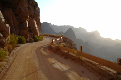 A winding dirt road along rocky cliffs, with mountains in the background and a warm sunset glow.