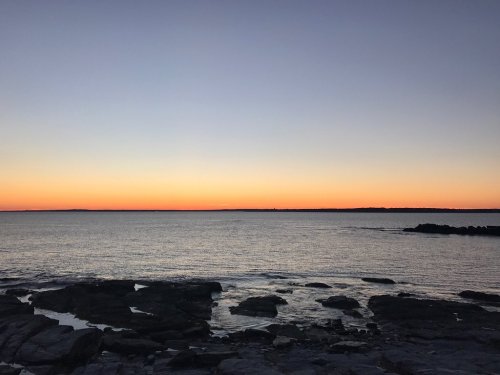 A serene seascape at sunset, with calm waters reflecting hues of orange and blue against a rocky shoreline.