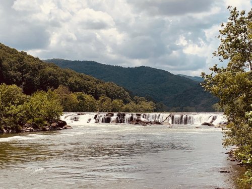 A serene river with cascading waterfalls, surrounded by lush green trees and mountains under a cloudy sky.