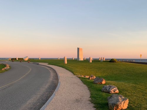 A serene landscape at sunset featuring a winding path, grassy area, and abstract sculptures near the ocean.