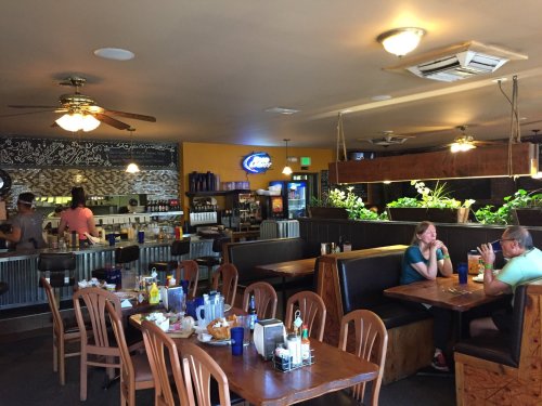Interior of a casual restaurant with wooden tables, a bar area, and patrons dining and chatting. Green plants visible.
