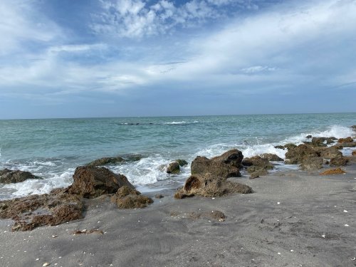 A serene beach scene with gentle waves lapping against rocky shore under a partly cloudy sky.