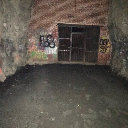A dimly lit cave with brick walls, featuring a barred door and graffiti on the rocks. The ground is dark and uneven.
