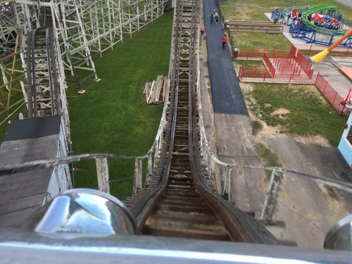 View from the top of a wooden roller coaster, showing the steep drop and surrounding amusement park area.