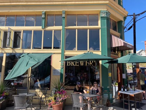 A brewery with large windows, outdoor seating, and green umbrellas on a sunny day. People are enjoying drinks outside.