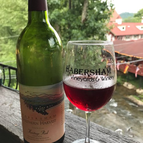 A bottle of Habersham Southern Harvest wine next to a glass of red wine, with a scenic background of greenery and buildings.