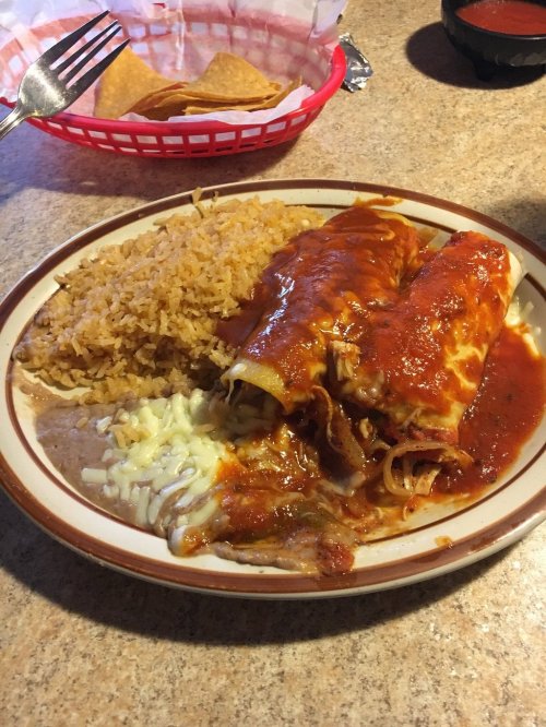 A plate of enchiladas topped with red sauce, served with rice and refried beans. Chips and salsa in the background.