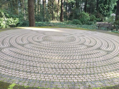 A circular stone labyrinth set in a forested area, surrounded by greenery and a bench in the background.