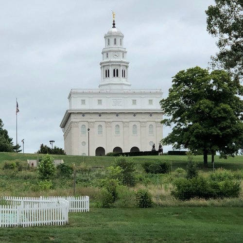 A white historic building with a tall steeple, surrounded by greenery and a cloudy sky.