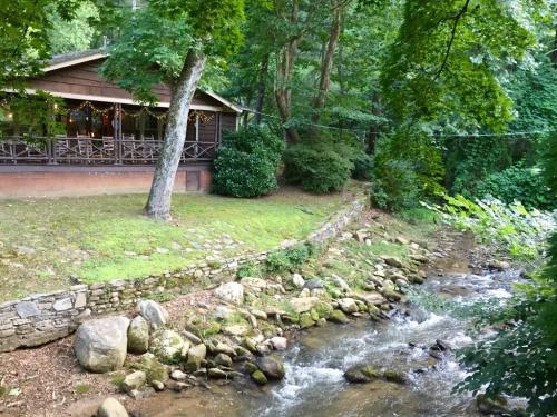 A cozy cabin by a stream, surrounded by lush greenery and trees, with a stone path leading to the water.