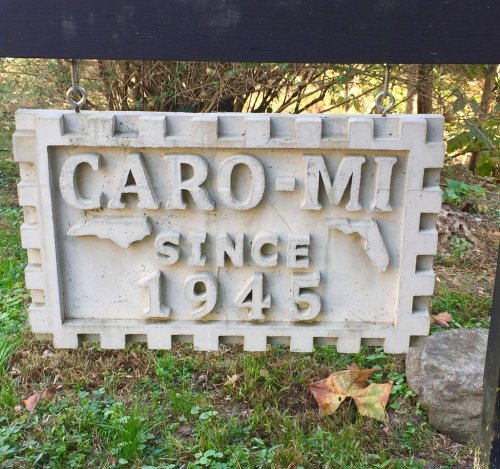 A stone sign reading "CARO-MI" with the text "SINCE 1945," surrounded by grass and trees.