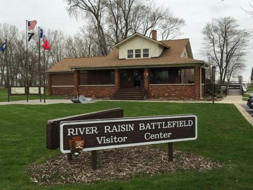 A brick visitor center with a sign reading "River Raisin Battlefield" surrounded by trees and flags.