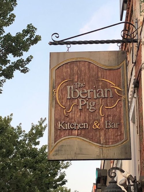 Sign for "The Iberian Pig Kitchen & Bar" hanging from a building, surrounded by trees and a clear sky.