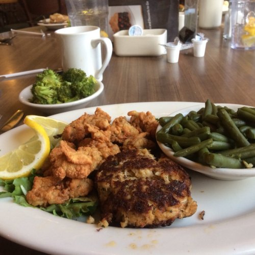 A plate of fried shrimp, a crab cake, green beans, and broccoli, garnished with lemon slices on a wooden table.