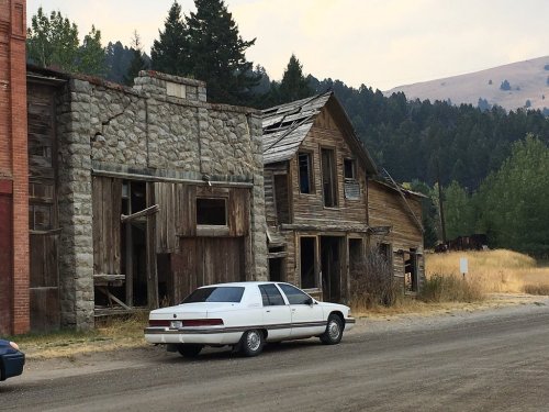 Abandoned buildings with weathered wood and stone, a vintage car parked on a dirt road surrounded by trees and hills.