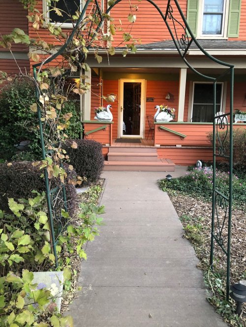 A pathway leads to a warmly lit entrance of an orange house, flanked by decorative swan statues.