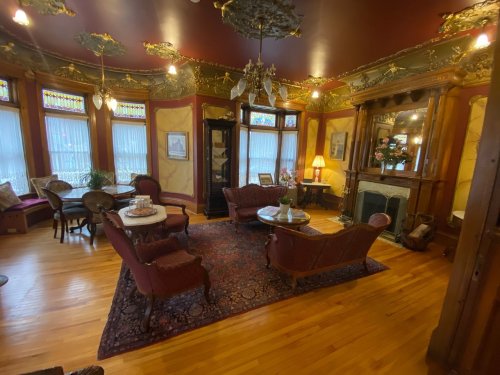 A vintage living room with ornate decor, wooden furniture, a fireplace, and large windows with stained glass.