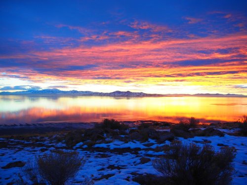 Vibrant sunset over a calm lake, reflecting colorful clouds and snow-covered ground in the foreground.