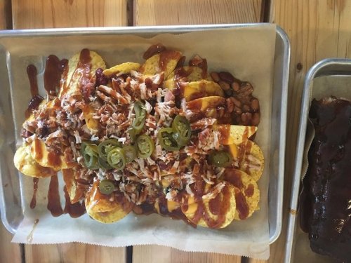 A tray of loaded nachos topped with pulled pork, cheese, jalapeños, beans, and barbecue sauce on a wooden table.