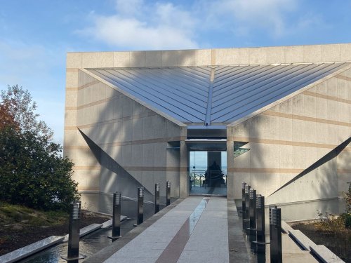 Modern building entrance with angular design, surrounded by greenery and a pathway leading to the door.