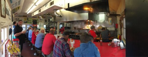 A bustling diner interior with patrons at the counter and cooks preparing food in the kitchen. Bright and lively atmosphere.