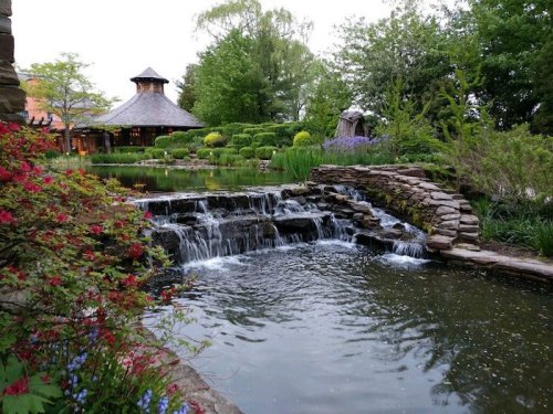 A serene garden scene featuring a waterfall, pond, and lush greenery with a gazebo in the background.