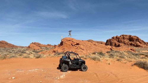 A person jumps on a rock in a desert landscape, with a black off-road vehicle parked nearby and red rock formations in the background.