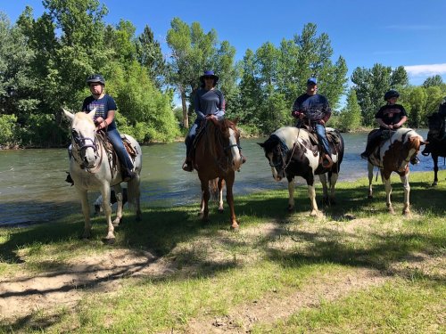 Four people on horseback stand by a river, surrounded by trees and a clear blue sky.