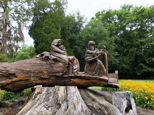 Wooden sculpture of three figures seated on a log, surrounded by greenery and yellow flowers in a park setting.