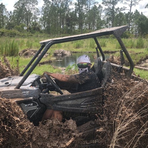 A person driving an off-road vehicle splashes through muddy water in a wooded area.