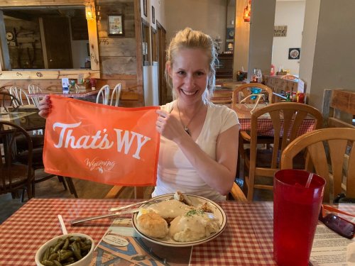 A smiling woman holds an orange "That's WY" flag in a cozy restaurant, with a plate of food in front of her.