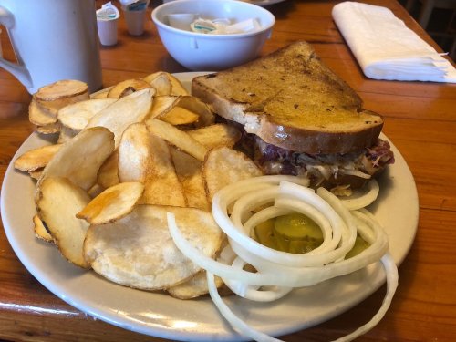 A plate with a sandwich, crispy potato chips, sliced onions, and pickles, alongside a cup and bowl in a casual dining setting.
