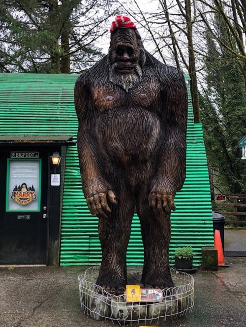 A large wooden Bigfoot statue wearing a red hat stands outside a green building, surrounded by a wire fence.