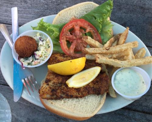 A plate with a fried fish sandwich, lettuce, tomato, cole slaw, fries, and a lemon wedge on a wooden table.