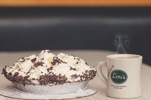 A slice of chocolate cream pie topped with whipped cream, next to a steaming cup of coffee on a table.