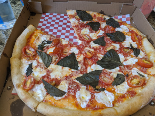 A pizza with fresh basil, cherry tomatoes, and ricotta cheese, partially eaten, in a pizza box with red checkered paper.