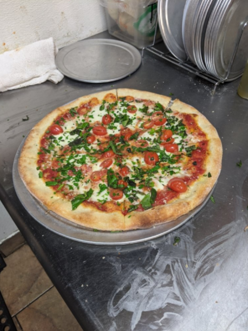 A freshly baked pizza topped with tomatoes, herbs, and melted cheese on a metal tray in a kitchen setting.