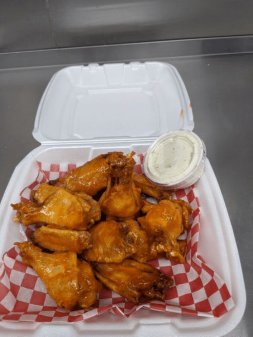 A takeout container filled with buffalo chicken wings and a side of ranch dressing on a checkered paper liner.