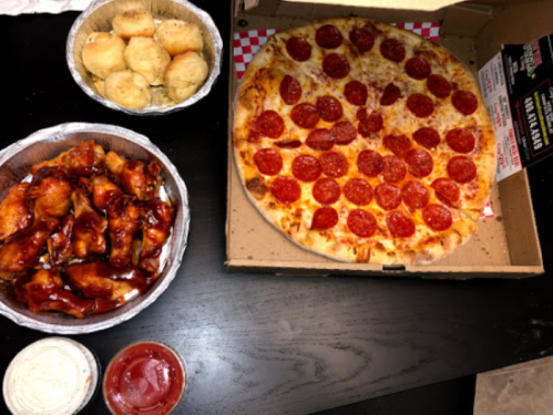 A table with a large pepperoni pizza, chicken wings in barbecue sauce, and two bowls of breadsticks with dipping sauces.